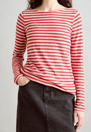 Chandail rayé rouge et crème à manches longues avec un col bateau, porté avec une jupe en denim noire. Présente une coupe décontractée et une main visible dans la poche.