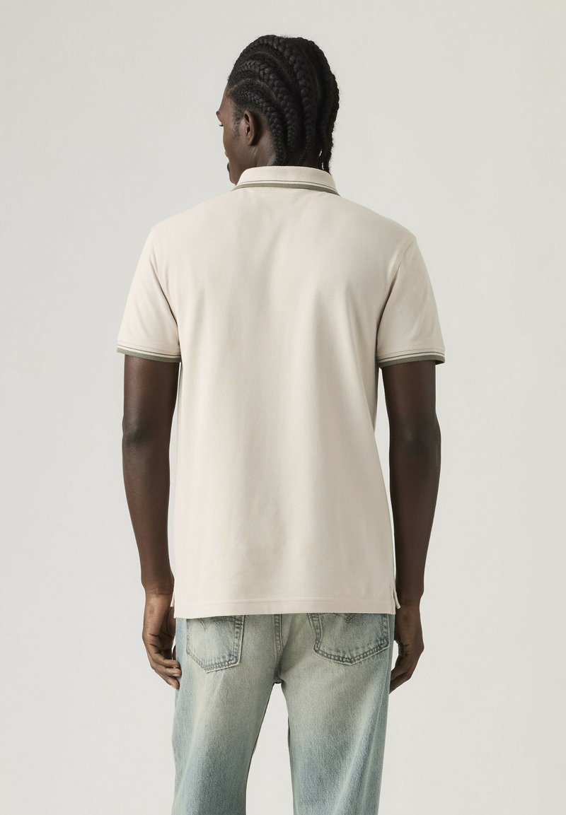 Homme aux cheveux tressés portant un polo à manches courtes beige clair et un jean délavé bleu clair, vu de dos sur un fond uni.