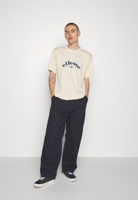 Camiseta de algodón beige con logo azul marino, combinada con pantalones de pierna ancha azul marino. Las zapatillas presentan una mezcla de colores verde, azul marino y blanco.