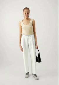 Débardeur beige en tricot, pantalons blancs à jambes larges avec des rayures verticales, baskets grises et blanches, tenant un petit sac à main noir.