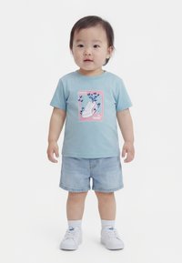 Småbarn iført lyseblå t-shirt med grafik af sneakers og blomster, denimshorts, hvide sokker og hvide Puma-sneakers, stående med fronten vendt fremad.