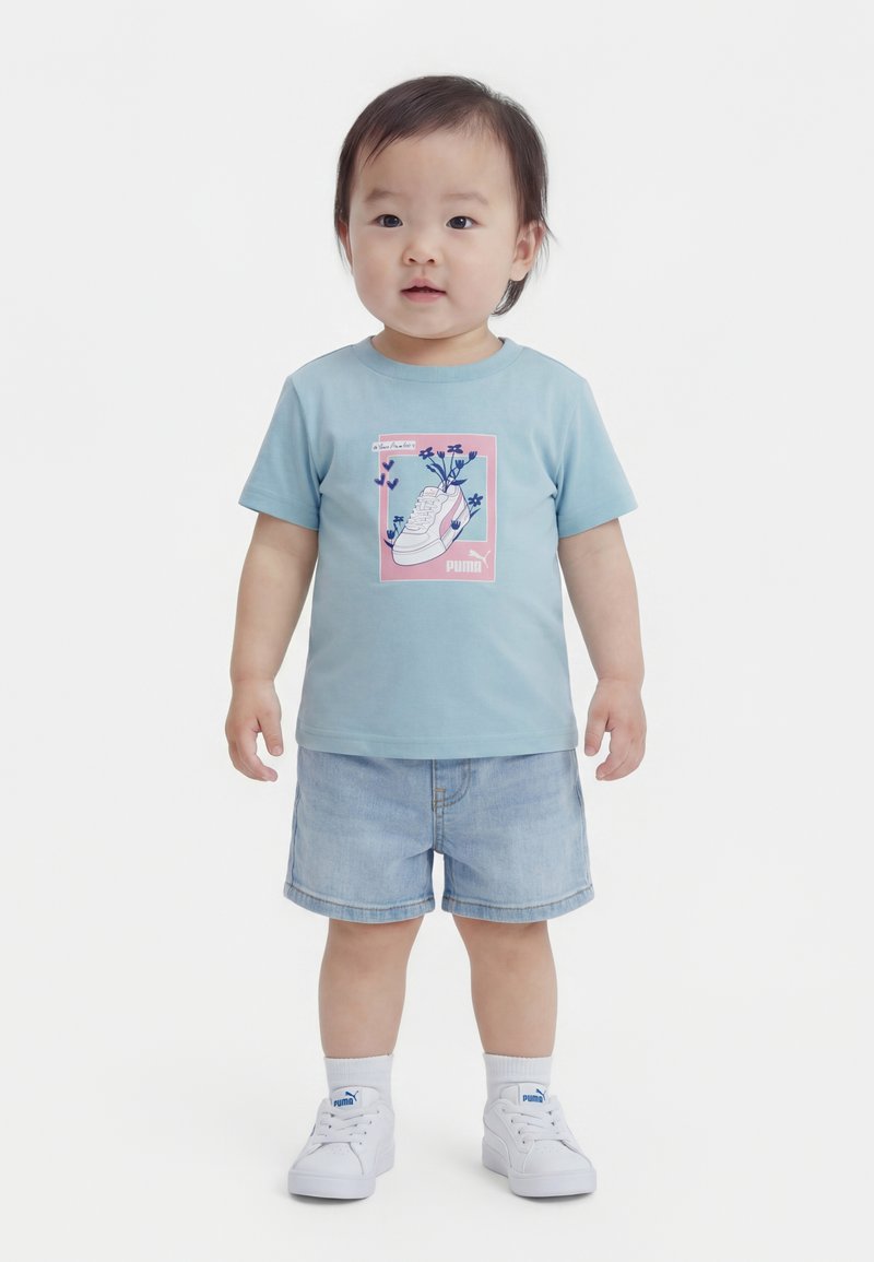 Småbarn iført lyseblå t-shirt med grafik af sneakers og blomster, denimshorts, hvide sokker og hvide Puma-sneakers, stående med fronten vendt fremad.
