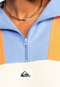 Pullover zip-up blu, arancione e bianco realizzato in tessuto testurizzato, con una toppa logo e una cerniera azzurra chiara con colletto angolare.