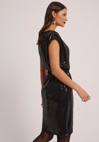 Czarna sukienka z cekinami o dopasowanej sylwetce, z krótkimi rękawami i wcięciem w talii. Błyszcząca tekstura i długość do kolan.