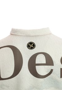 Camisa blanca texturizada con un pequeño parche de logo negro y dorado en el cuello de la parte posterior y letras grandes marrones en la tela.