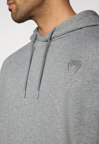 Γκρι φούτερ με μαλακή υφή, με κουκούλα με κορδόνι, ανοιχτογκρι λεπτομέρειες και διακριτικό λογότυπο στο στήθος.