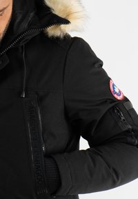 Parka noire avec capuche en fausse fourrure, fermeture éclair et patch de marque sur la manche. Présente des poignets côtelés et des poches latérales.