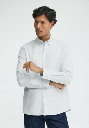 Uomo con capelli scuri che indossa una camicia bianca a maniche lunghe con bottoni e pantaloni scuri, in piedi con le braccia incrociate davanti a uno sfondo semplice.