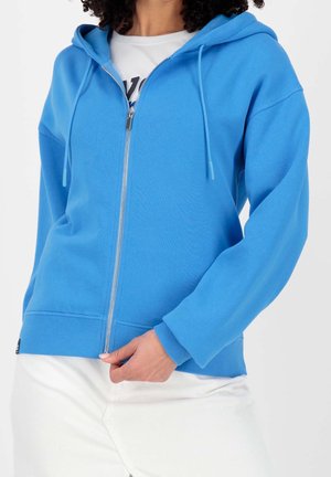 Femme portant un sweat à capuche bleu vif zippé sur un t-shirt blanc, associé à un pantalon blanc, tenant la tirette de la fermeture éclair près de la taille.