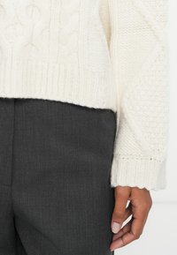 Suéter blanco de punto trenzado con patrones texturizados y dobladillo acanalado, combinado con pantalones oscuros de corte entallado. La mano reposa suavemente sobre la tela.