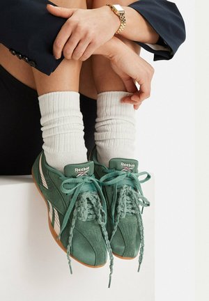 Osoba nosząca białe skarpetki i zielone buty Reebok, siedząca z założonymi na siebie ramionami na zgiętych kolanach, ubrana w złoty zegarek na nadgarstku i ciemne ubrania.