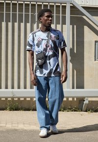 Grafická tričko v modrej a čiernej farbe s ružovými akcentmi, kombinované s voľnými modrými džínsy a bielymi teniskami. Obsahuje malú čiernu tašku cez telo.