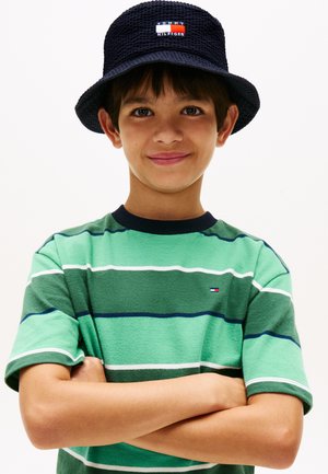 Αγόρι με μπλε καπέλο τύπου κουβά και πράσινο ριγωτό πουκάμισο στέκεται με σταυρωμένα τα χέρια, χαμογελώντας με αυτοπεποίθηση μπροστά σε απλό φόντο.