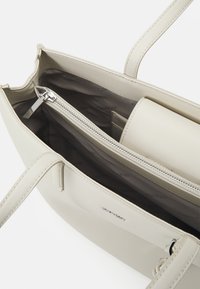 Borsa a tracolla in pelle beige chiaro con texture liscia, chiusura con zip superiore e un interno compartimentato che include una piccola tasca.