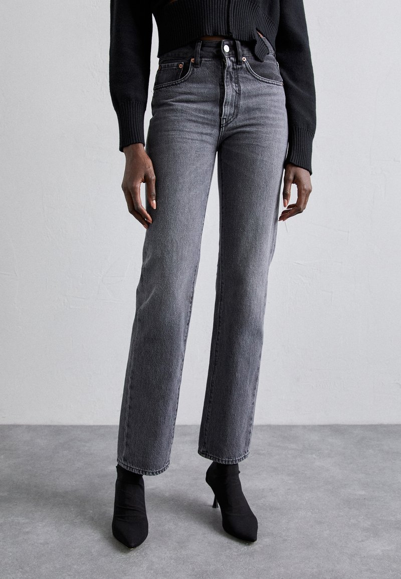 High-waisted, straight-leg black jeans with a faded wash, paired with black ankle boots. Material is denim; features classic five-pocket design.