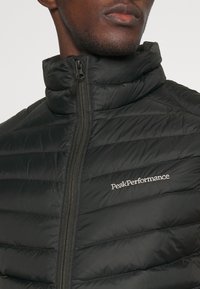Czarna pikowana kurtka puchowa z wysokim kołnierzem i pełnym zamkiem, z dyskretnym białym logo "PeakPerformance" na piersi.