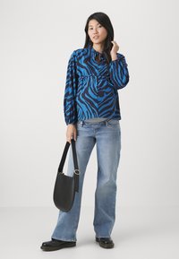 Blå og svart zebra-stripete bluse med lange puffermer og nøkkelhullsår, kombinert med lyseblå jeans og en svart skulderveske.
