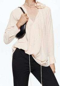 Helle Pfirsichfarbene Bluse mit langen Ärmeln, V-Ausschnitt, fließendem Stoff und strukturiertem Blumenmuster auf der Schulter, kombiniert mit schwarzen Hosen und einer kleinen Tasche.