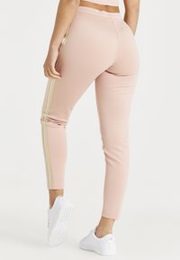 Růžové sportovní kalhoty se zlatými bočními pruhy, které mají přiléhavý střih a elastický pás, doplněné bílými teniskami.