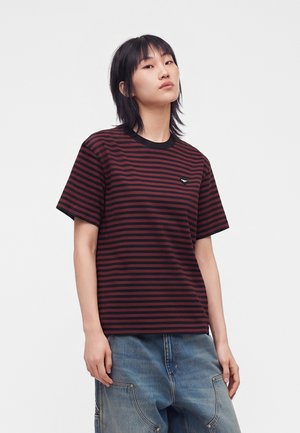 Ung person med glat mørkt hår iført en sort og mørkerød stribet t-shirt og løse blå jeans, stående mod en ensfarvet baggrund.