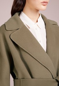 Manteau texturé vert olive avec un large revers, un design enroulé et une ceinture, porté par-dessus une chemise blanche à col avec des boutons visibles.