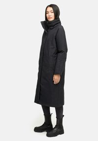 Kvinde i en lang sort polstret frakke med høj krave, sorte skinny jeans og sorte klodsede ankelstøvler, som står sidelæns.