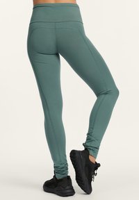 Kvinna som bär högmidjade turkosa leggings och svarta träningsskor, stående med det ena benet lätt böjt och hälen upphöjd.