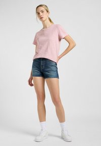 Różowa koszulka z bawełny slubowej z krótkim rękawem, w zestawie z niebieskimi szortami dżinsowymi. Model nosi białe trampki i skarpetki za kostkę. Prosty, casualowy strój.