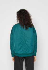 Teal bomberjakke med en glat tekstur, ribbede manchetter og talje. Har quilted detaljer på ærmerne og et enkelt design på ryggen.