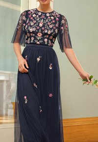 Marineblaues Kleid mit floralem Stickerei auf dem Oberteil und dem Rock. Durchsichtige Ärmel und fließender Stoff. Model hält einen kleinen Blumenstrauß.