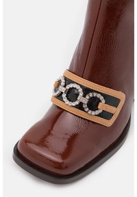 Bottine en cuir verni marron avec un bout carré, dotée d'une lanière décorative noire et beige ornée de pierres précieuses argentées et noires.