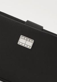 Fekete tárca textúrált anyagból, fényes ezüst téglalap alakú logó táblával, rajta "TOMMY JEANS" felirattal. Kompakt és minimalista dizájn.