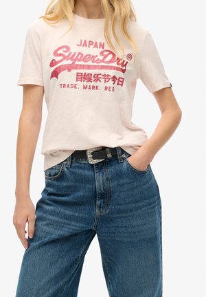 Camiseta de manga corta color rosa claro con gráfico rojo "SuperDry". Combinada con jeans de mezclilla azul de talle alto y un cinturón negro con hebilla metálica.