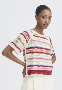 Žena s účesom stáhnutým do copu, nosiaca náušnice v tvare kruhu a farebný háčkovaný top s krátkym rukávom, pózujúca s rukami na bokoch.