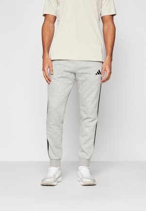 Mężczyzna w jasnobeżowym T-shircie, jasnoszarych dresach Adidas z czarnymi paskami i białych trampkach, stoi na jednolitym tle.