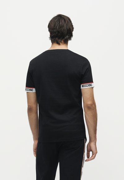 Homme aux cheveux foncés portant un t-shirt et un pantalon noirs Moschino, vu de dos sur un fond clair uni.