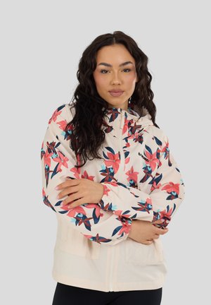 Frau mit lockigem dunklem Haar, die eine weiße Jacke mit rosa-blauem Blumenmuster trägt und mit verschränkten Armen vor einem grauen Hintergrund steht.