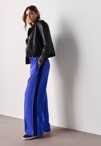 Fekete bőr motoros dzseki, testhez simuló fazonban. Laza kék melegítőnadrág fekete oldalsávokkal. A modell semleges háttér előtt pózol.