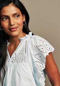 Blusa azzurra chiara con delicati dettagli in ricamo a occhio di pernice, scollo a volant e maniche corte. Morbida al tatto, vestibilità comoda.