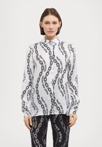 Λευκό πουκάμισο με κουμπιά και σχέδιο από μαύρη αλυσίδα και κείμενο; μακριά μανίκια; κλασικό γιακά; συνδυασμένο με μαύρες παντελονιές με σχέδιο.