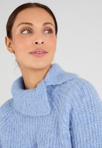 Suéter de punto azul claro con un cuello alto, que presenta una textura suave y esponjosa y puntos sueltos. Primer plano del escote y los hombros.