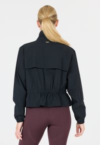 Black cropped jacket with a ruffled hem, elastic cuffs, and high collar; features a contrasting tag at the back. Smooth, lightweight fabric.