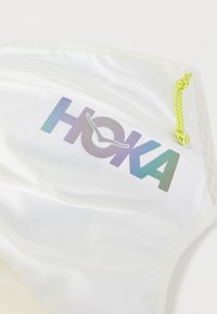 Λευκό ύφασμα με αντανακλαστικό λογότυπο HOKA σε διαβαθμισμένα χρώματα και κίτρινο κορδόνι δεμένο σε κόμπο στο πάνω δεξί μέρος.