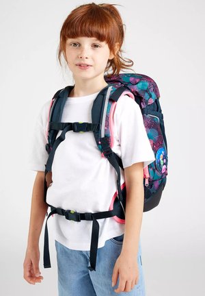 Junges Mädchen mit roten Haaren, das ein weißes T-Shirt und blaue Jeans trägt und einen bunten Rucksack mit Blumenmustern und schwarzen Sicherungsgurten auf dem Rücken hat.