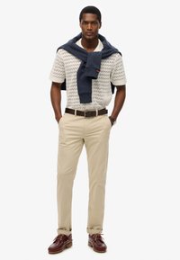 Chinos beige y una camisa de punto blanca con estampado, combinados con un suéter azul marino. Unos zapatos de barco de cuero marrón completan el look.