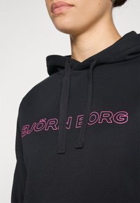Sweatshirt preto com um bolso frontal, com o texto ousado em rosa "BJÖRN BORG." O material parece suave, com um típico cordão na capuz.