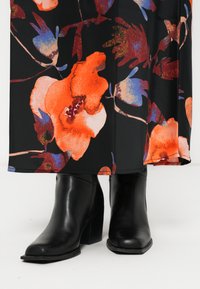 Zwarte leren enkellaarsjes met een vierkante teen en robuuste hak, gecombineerd met een zwarte bloemenjurk met levendige oranje en blauwe patronen.