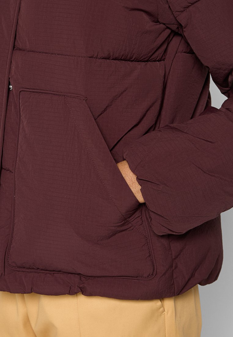 Giacca a vento bordeaux con finitura texturizzata, dotata di tasca laterale e polsini elasticizzati per una vestibilità aderente. Il materiale sembra leggero.