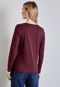 Bordeauxfarbenes Langarmoberteil aus weichem Stoff mit Rundhalsausschnitt und lässiger Passform, kombiniert mit dunkelblauen Jeans. Glatte Textur, minimalistische Details.