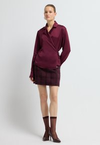 Bordeaux satin omslagbluse med lange ærmer, parret med en ternet miniskirt i komplementerende nuancer, afsluttet med høje hæle og gennemskinnelige sokker.